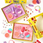 Two open yellow boxes containing the Super Smalls Rainbow Gems Beauty Play Kit—a kid-safe makeup set featuring lipsticks, blush, brushes, nail decals, and jewelry—are displayed on a white background. Cosmetic items from the kit are scattered around the boxes, making them perfect for pretend play fun.