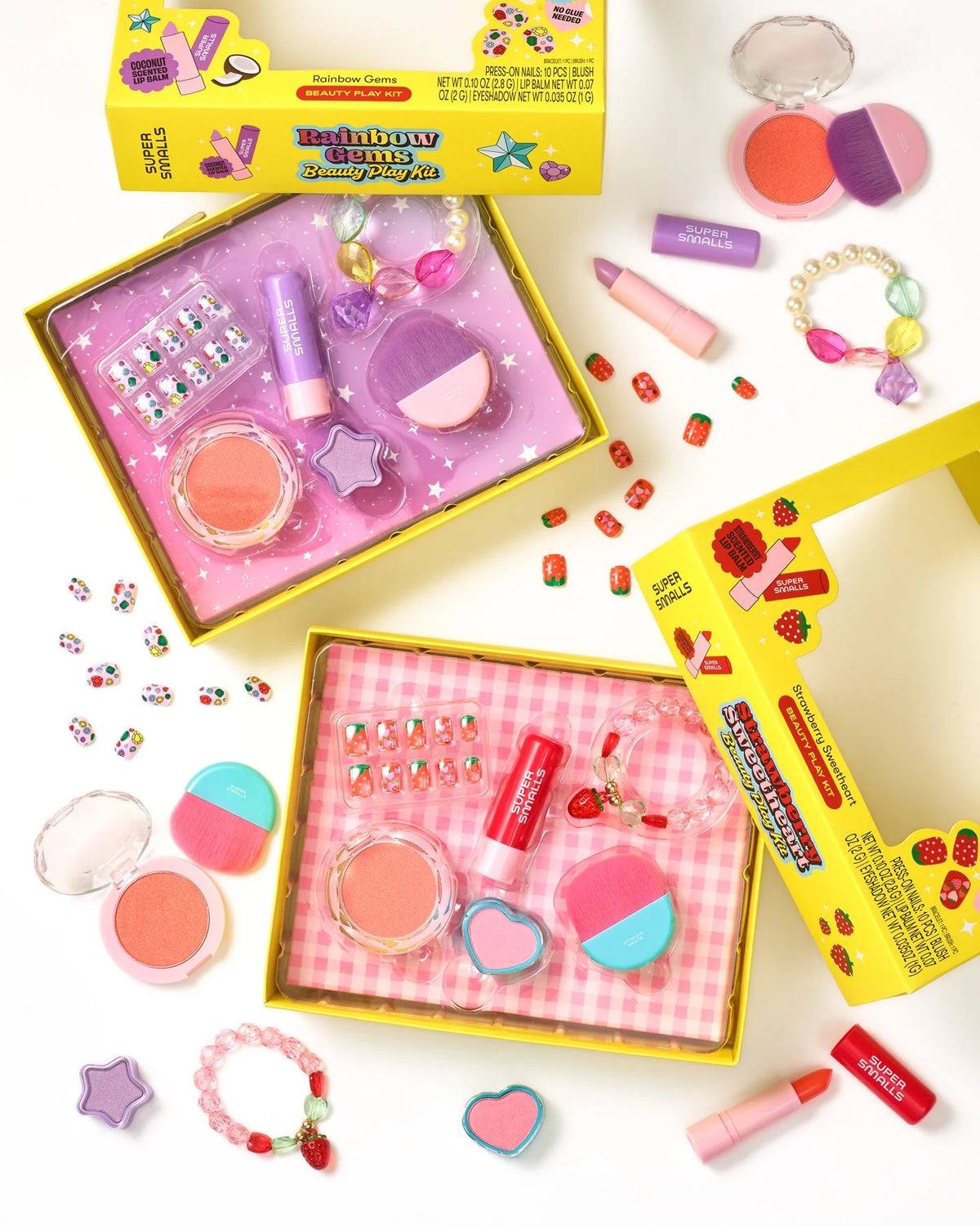 Two open yellow boxes containing the Super Smalls Rainbow Gems Beauty Play Kit—a kid-safe makeup set featuring lipsticks, blush, brushes, nail decals, and jewelry—are displayed on a white background. Cosmetic items from the kit are scattered around the boxes, making them perfect for pretend play fun.