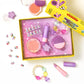 Box with colorful kids’ pretend makeup, including lip balm, blush, brushes, star-shaped containers, play rings, and a beaded bracelet on a white background—perfect for pretend play. The Strawberry Sweetheart Beauty Play Kit by Super Smalls features kid-safe makeup for hours of imaginative fun.