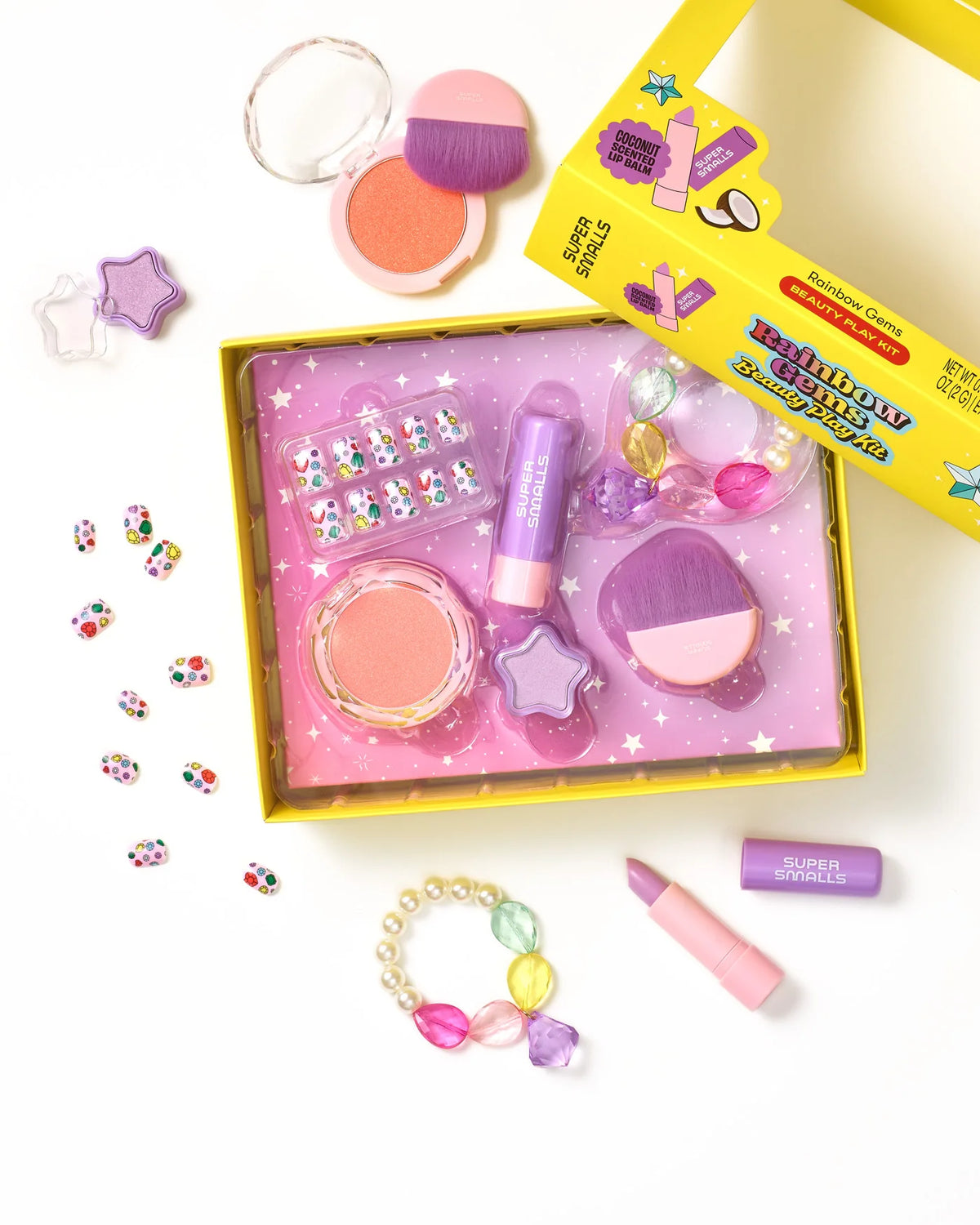 Box with colorful kids’ pretend makeup, including lip balm, blush, brushes, star-shaped containers, play rings, and a beaded bracelet on a white background—perfect for pretend play. The Strawberry Sweetheart Beauty Play Kit by Super Smalls features kid-safe makeup for hours of imaginative fun.