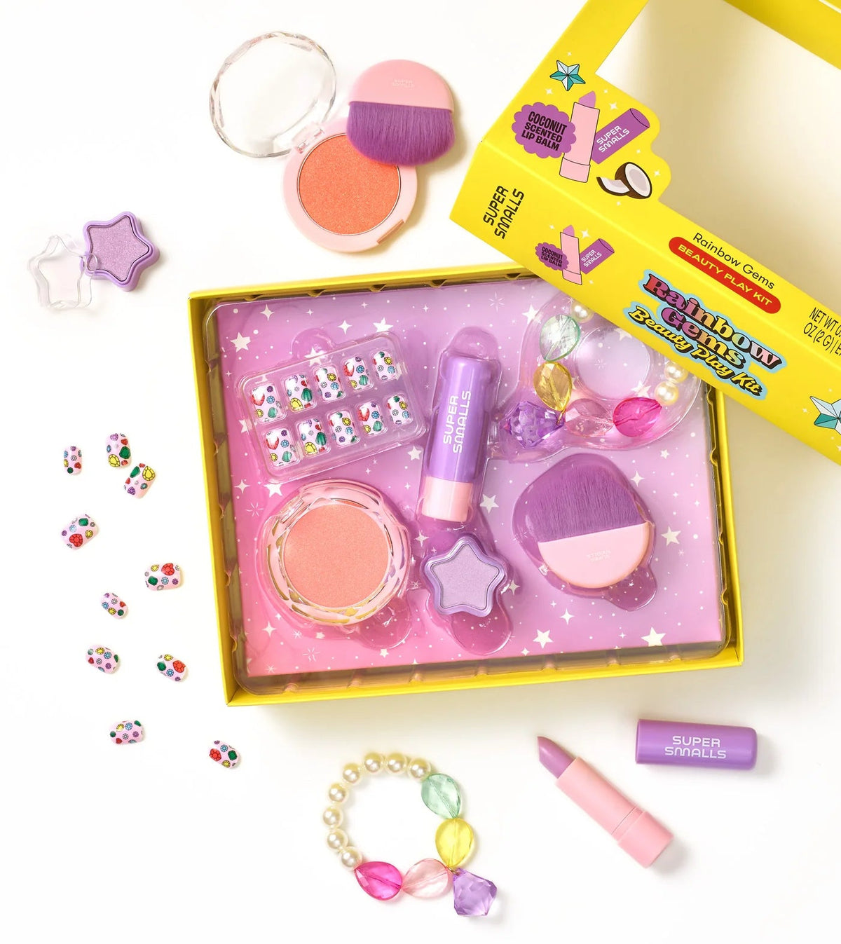 A yellow box contains the Super Smalls Rainbow Gems Beauty Play Kit, which includes kid-safe makeup for pretend play, toy brushes, pretend lipstick, colorful beads, rings, and stick-on nails, all set against a purple star-patterned background.