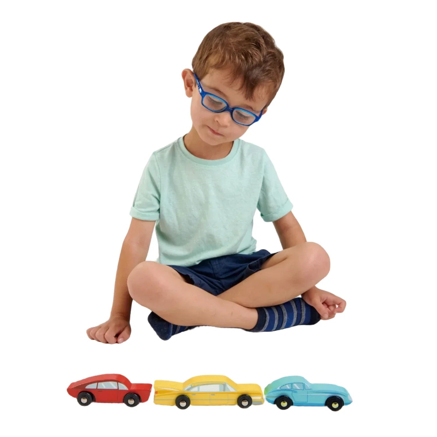 A young boy with glasses sits cross-legged on the floor, looking down at three Retro Cars by Tender Leaf Toys—red, yellow, and blue—lined up in front of him.