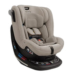 The Nuna Revv Carseat by Nuna is a gray convertible car seat with a black base, featuring a harness, headrest, and recline adjustments. This rotating car seat includes side impact protection, various safety labels, and an instructional panel on the base for simplifying rear to forward-facing transitions.