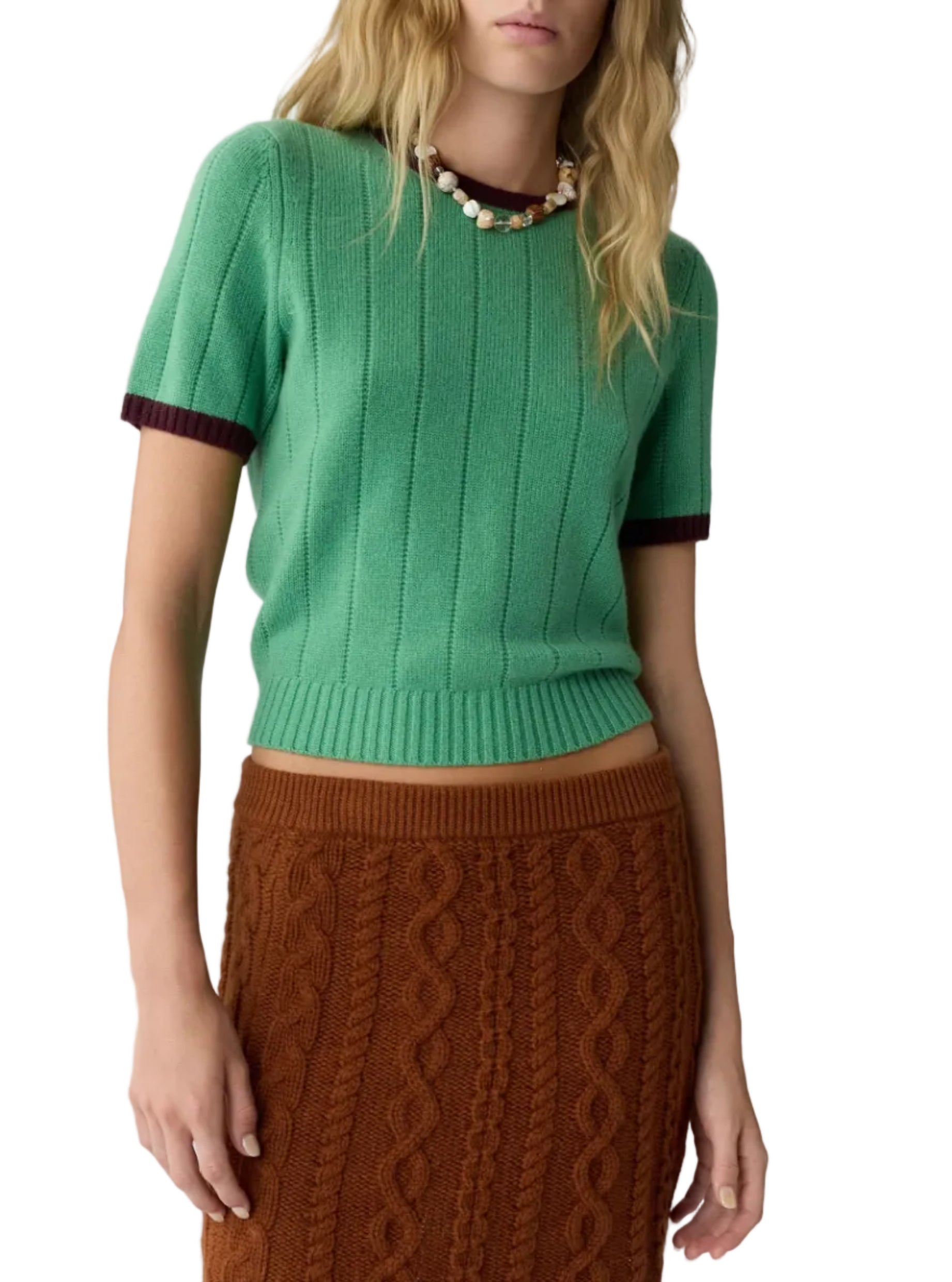 A woman wears the Guest in Residence Rib Ringer Tee, a green women’s cashmere knit short-sleeve sweater with maroon sleeve trim, paired with a brown cable knit skirt and accessorized with a beaded necklace.
