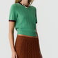 A woman wearing the Guest in Residence Rib Ringer Tee—a green women’s cashmere knit short-sleeve sweater with maroon trim—paired with a brown cable knit skirt and a beaded necklace, posed against a plain background.