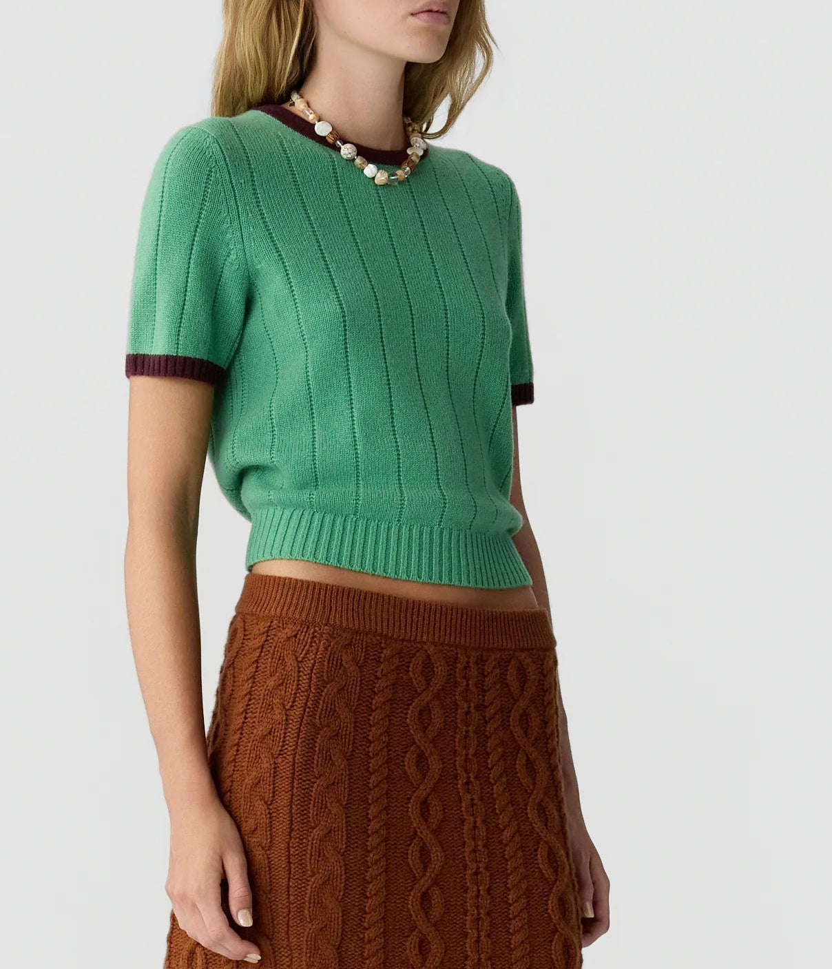 A woman wearing the Guest in Residence Rib Ringer Tee—a green women’s cashmere knit short-sleeve sweater with maroon trim—paired with a brown cable knit skirt and a beaded necklace, posed against a plain background.