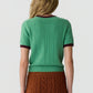 A woman faces away, wearing the Guest in Residence Rib Ringer Tee in green with maroon trim, paired with a rust-colored cable knit skirt.