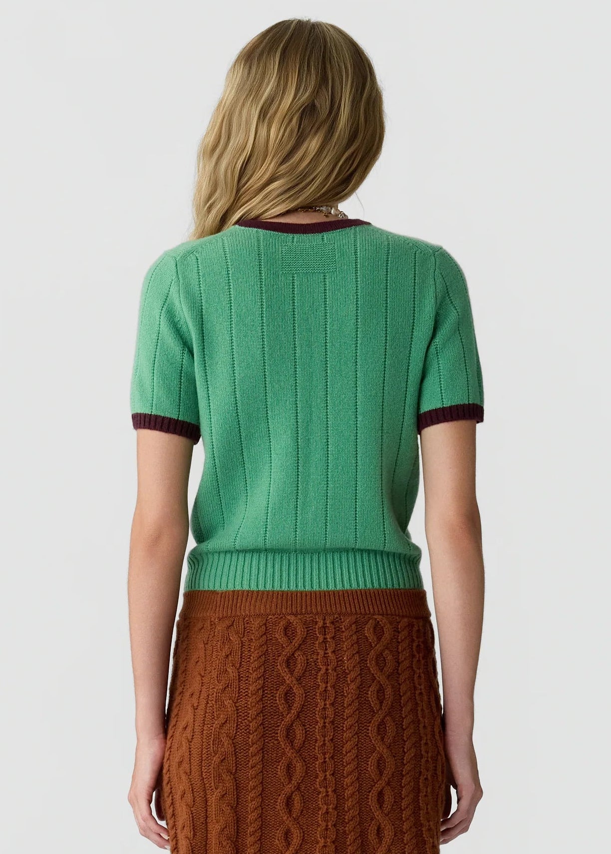 A woman faces away, wearing the Guest in Residence Rib Ringer Tee in green with maroon trim, paired with a rust-colored cable knit skirt.