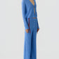A person stands against a plain background wearing Guest in Residence's Rib Everywear Pant and a matching blue ribbed knit cardigan with a front cutout, accessorized with a chunky necklace and sandals.