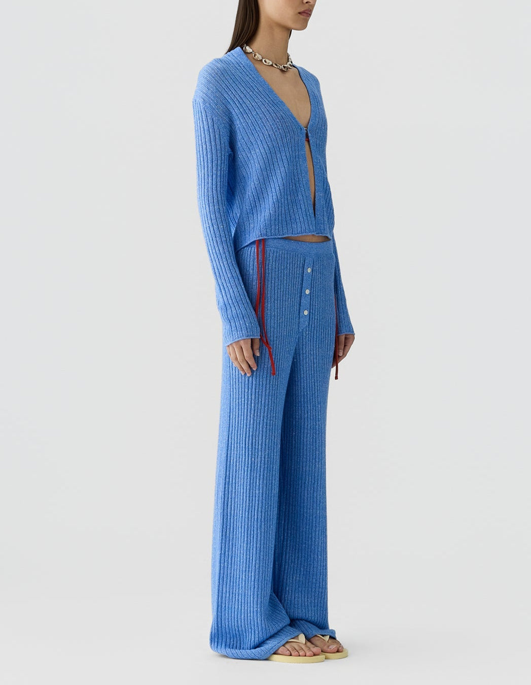 A person stands against a plain background wearing Guest in Residence's Rib Everywear Pant and a matching blue ribbed knit cardigan with a front cutout, accessorized with a chunky necklace and sandals.