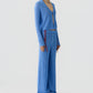 A person wears the Rib Stealth Cardigan by Guest in Residence with matching wide-leg pants featuring button details and sandals, standing against a plain light background.