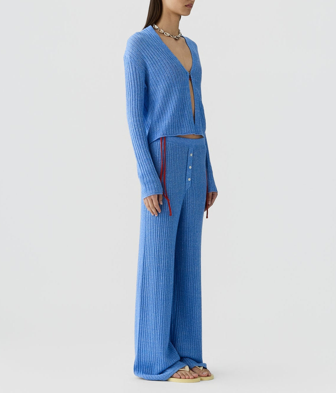 A person wears the Rib Stealth Cardigan by Guest in Residence with matching wide-leg pants featuring button details and sandals, standing against a plain light background.