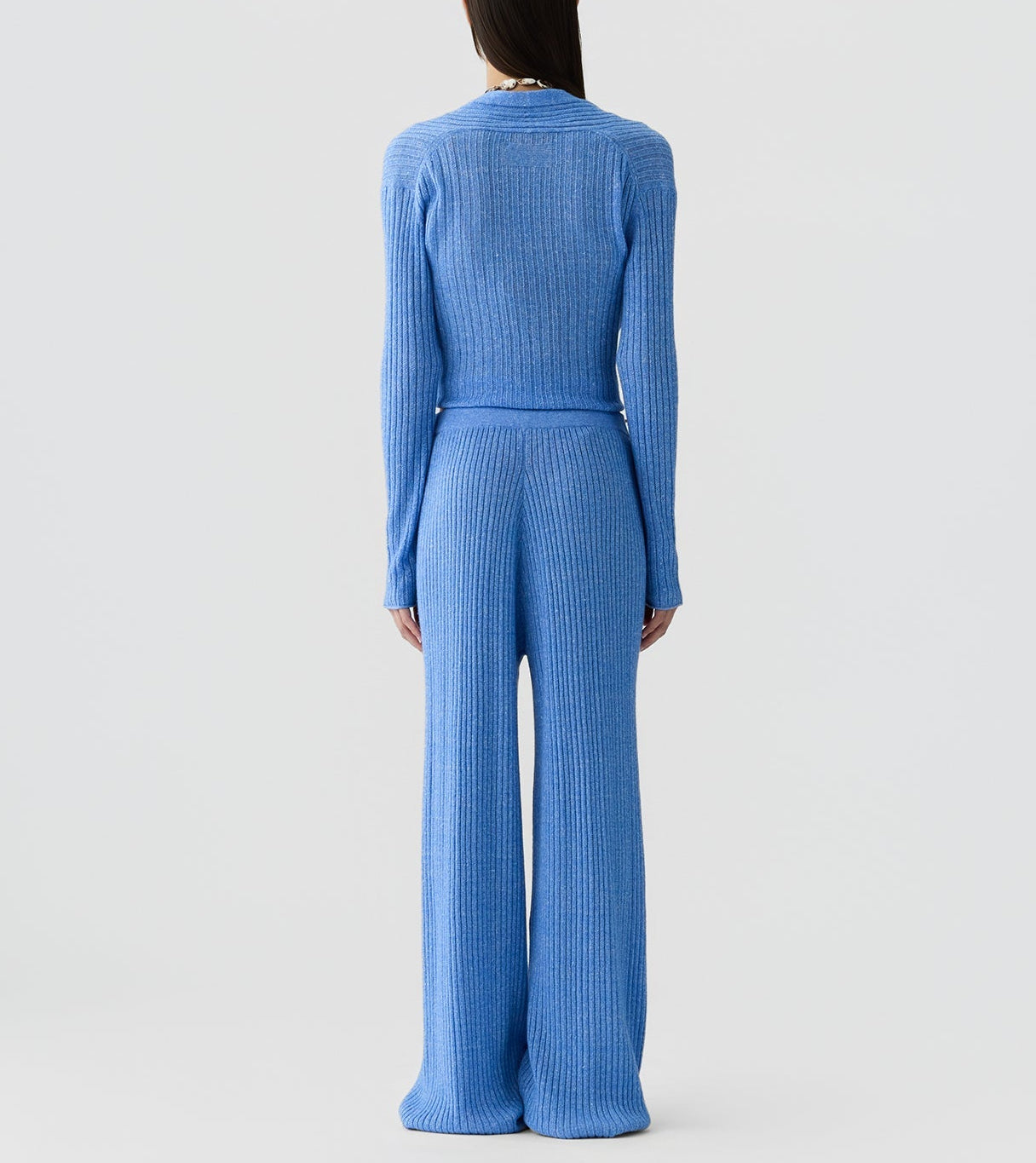 A person stands against a plain background, shown from the back, wearing Guest in Residence’s Rib Stealth Cardigan layered over a matching blue ribbed knit long-sleeve top and wide-leg pants.