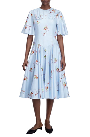A woman wears the Emilia Wickstead Risabelle Dress, a light blue, knee-length midi with short flutter sleeves and a delicate lily print, paired with black flats.