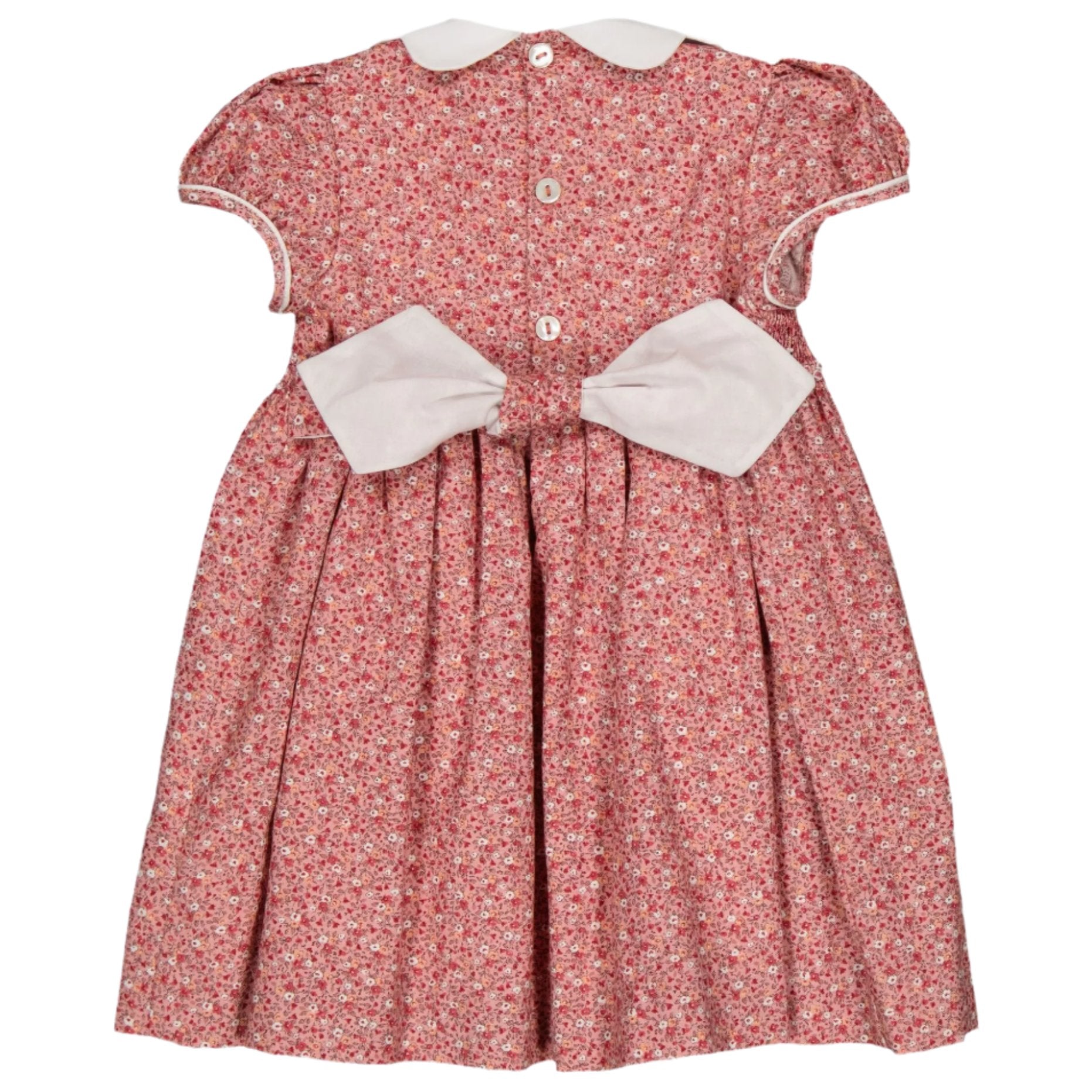 The Kidiwi Risette Smocked Dress is a petite red floral print dress featuring a white collar and cuffs, buttons along the back, and adorned with a prominent white bow.