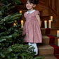 A young girl in a Kidiwi Risette Smocked Dress, crafted from organic cotton, stands on stairs beside a decorated Christmas tree and lit candles.