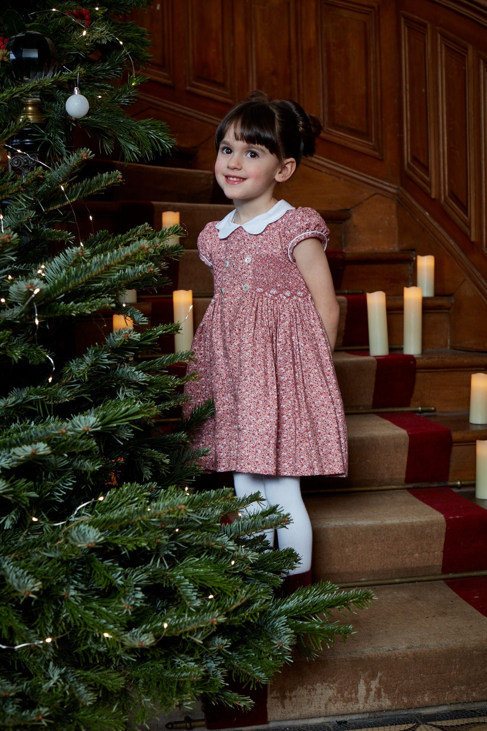 A young girl in a Kidiwi Risette Smocked Dress, crafted from organic cotton, stands on stairs beside a decorated Christmas tree and lit candles.