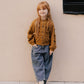 A young girl with long red hair models Rylee & Cru's Girls' Barrel Pant—loose blue pants with an elastic waistband—paired with a brown knit sweater and black shoes, standing by a beige wall with one hand in her pocket.