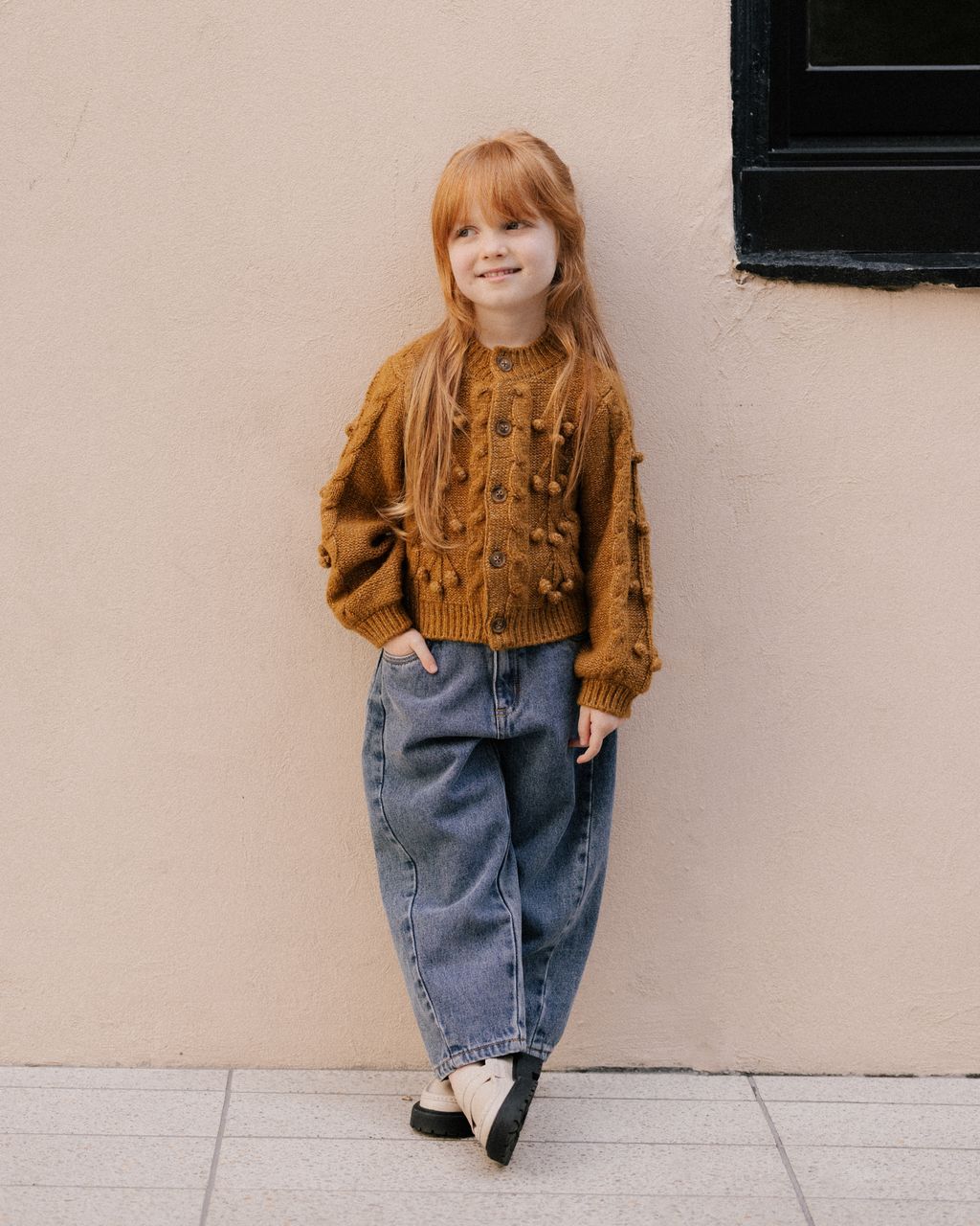 A young girl with long red hair models Rylee & Cru's Girls' Barrel Pant—loose blue pants with an elastic waistband—paired with a brown knit sweater and black shoes, standing by a beige wall with one hand in her pocket.