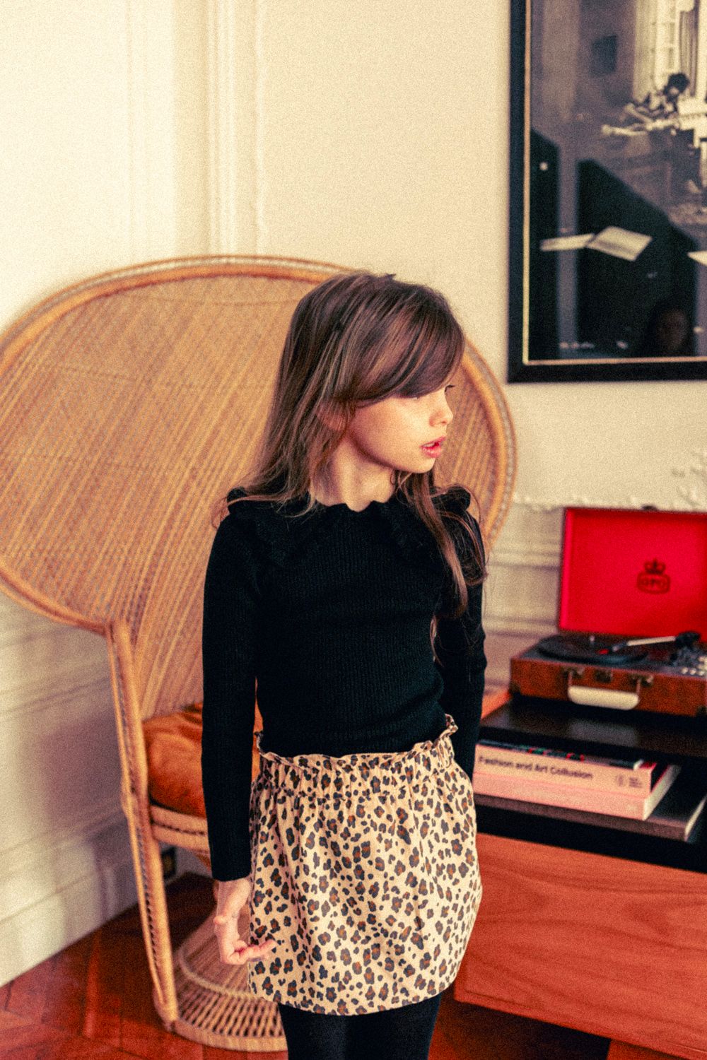 A young girl stands indoors wearing the Louis Louise Girls' Alinou Wool Blend Pull, paired with a leopard print skirt; a wicker chair and a red record player are in the background.