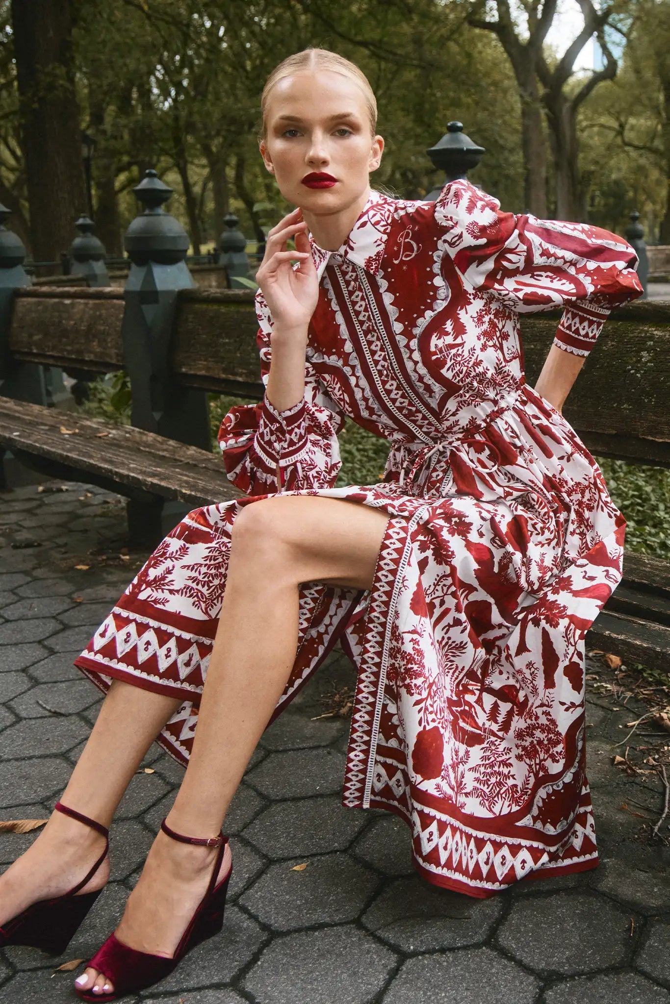 A person sits on a park bench wearing the Rosie Dress by Hunter Bell NYC with burgundy heels, surrounded by trees and a stone path—an elegant look that's perfect for holiday gatherings.