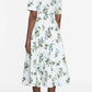 A person stands facing away, wearing the Rubie Dress by Emilia Wickstead—a white, short-sleeved cotton midi dress adorned with blue and green floral prints, perfect for summer—paired with black sandals against a plain background.