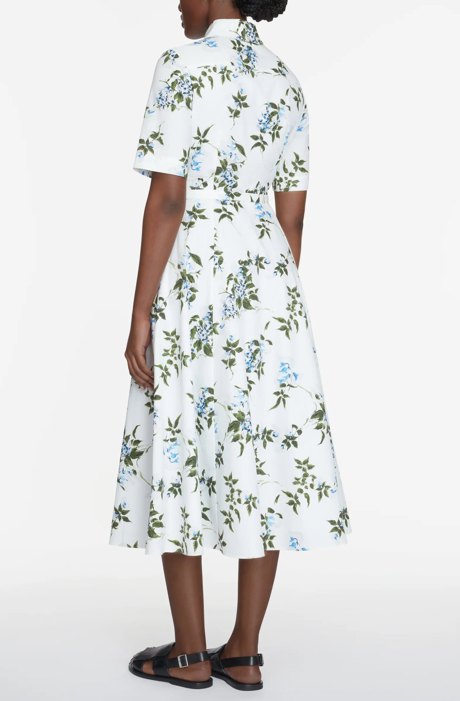 A person stands facing away, wearing the Rubie Dress by Emilia Wickstead—a white, short-sleeved cotton midi dress adorned with blue and green floral prints, perfect for summer—paired with black sandals against a plain background.