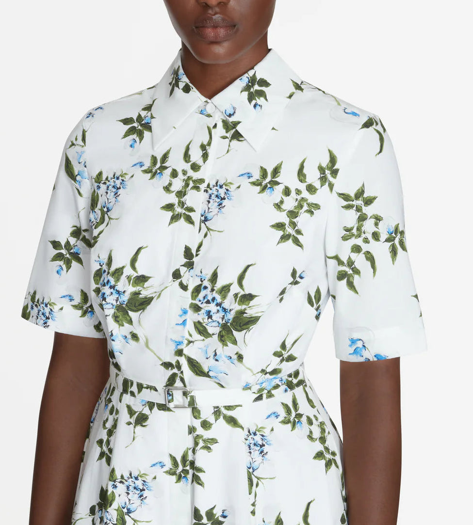 A person wearing the Rubie Dress by Emilia Wickstead, a white cotton dress with short sleeves featuring a green and blue floral print, shown from the shoulders to the waist.