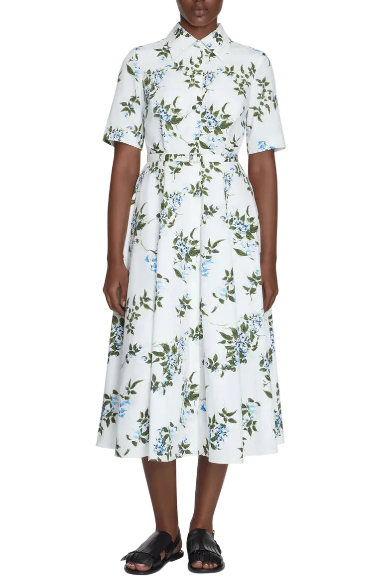 Wearing the Emilia Wickstead Rubie Dress, a white collared, short-sleeve button-down cotton dress adorned with a blue and green floral print, paired with black sandals, the person stands against a white background. The Rubie Dress effortlessly blends comfort and style for warm-weather days.