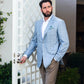 A bearded man wearing a Canali Sky Blue Fine Plaid Blazer, white shirt, and brown pants stands by a white lattice door, holding the handle.