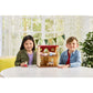 Two children sit at a table with the Calico Critters Red Roof Cozy Cottage and toy figures, including a Chocolate Rabbit girl, in front of a window decorated with white curtains and green pennants.