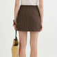 A woman stands facing away, wearing a white t-shirt, the POSSE Alice Pinstripe Skirt, strappy sandals, and holding a woven straw bag.