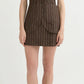 A woman wearing the POSSE Emma Pinstripe Vest, a brown sleeveless mini dress with a classic silhouette, front buttons, and pockets, stands against a plain background in flip-flops.