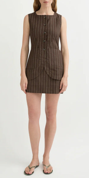 A woman wearing the POSSE Emma Pinstripe Vest, a brown sleeveless mini dress with a classic silhouette, front buttons, and pockets, stands against a plain background in flip-flops.