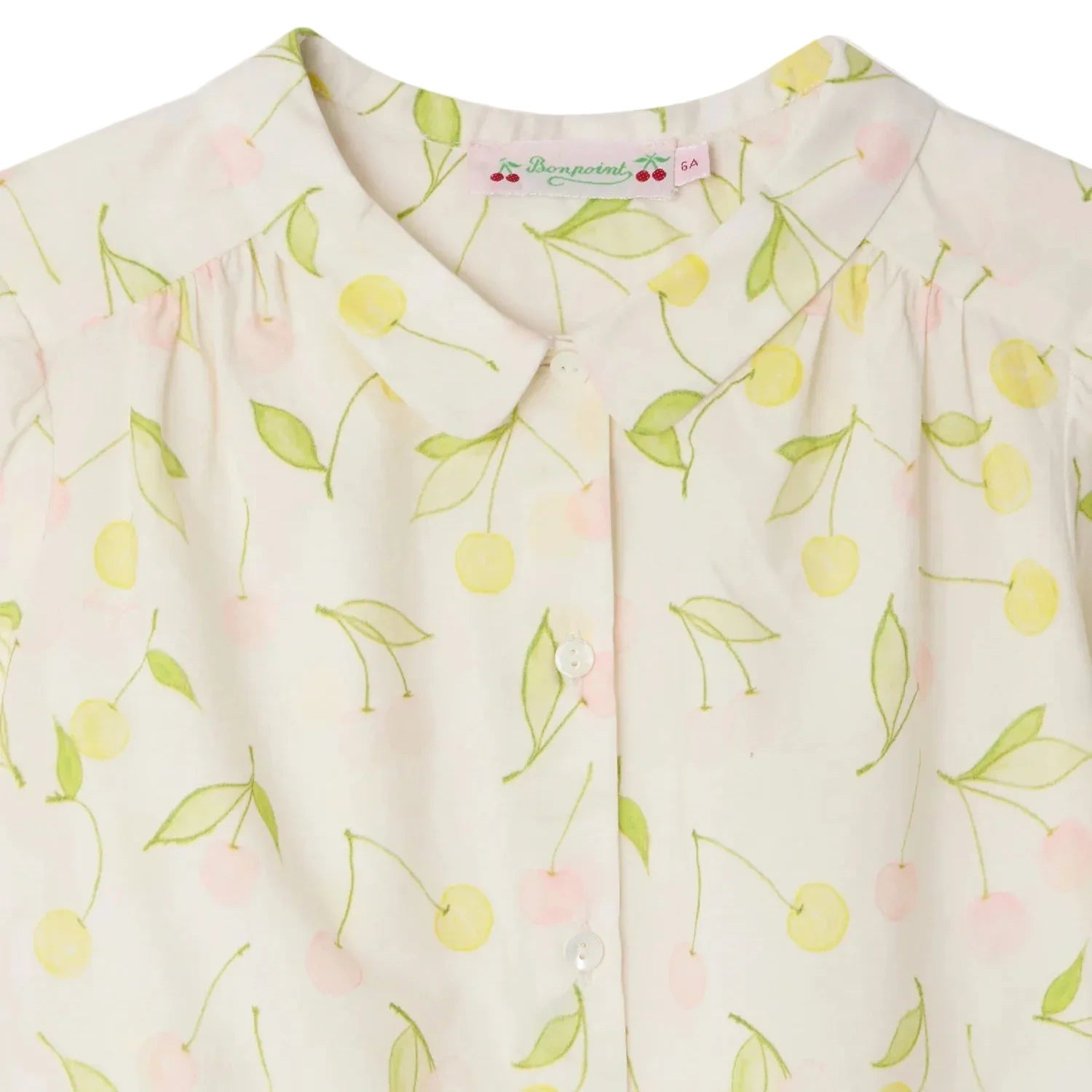 Close-up of Bonpoint's Girls' Adèle Blouse—crafted from light organic cotton, this blouse features a round collar, button front, and a cheerful cherry print in yellow and pink with green stems and leaves.