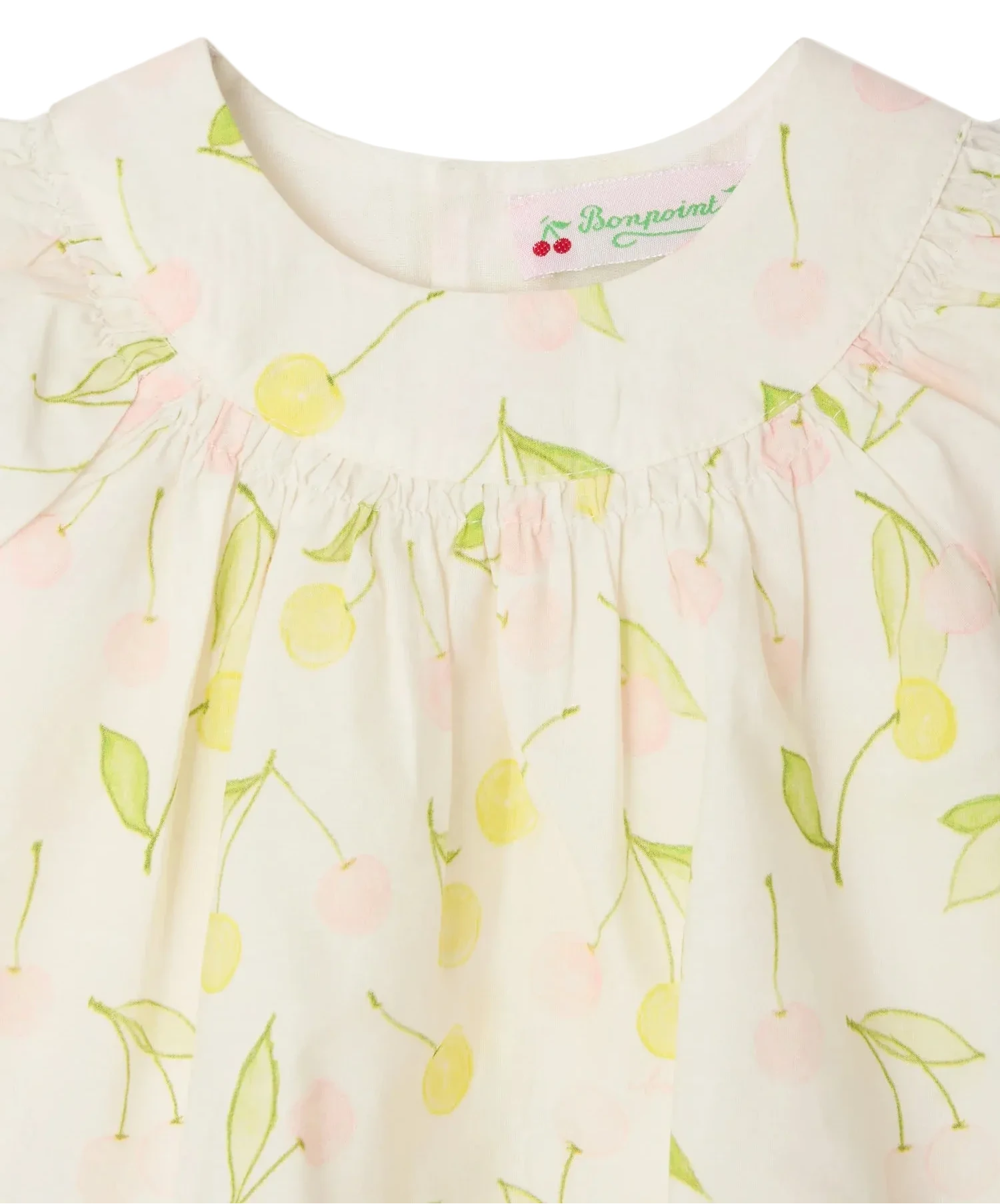 Close-up of the Bonpoint Baby Girls' and Girls' Lala Blouse in white organic cotton, with a yellow and pale pink cherry print, gathered neckline, mother-of-pearl buttons, and "Bonpoint" label inside the collar.