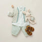 A neatly arranged baby outfit featuring a striped shirt, light green Bonpoint cardigan, mint Baby Boys' Dandy Trousers by Bonpoint, brown sandals, a plush bunny, and a wooden toy on a light background.