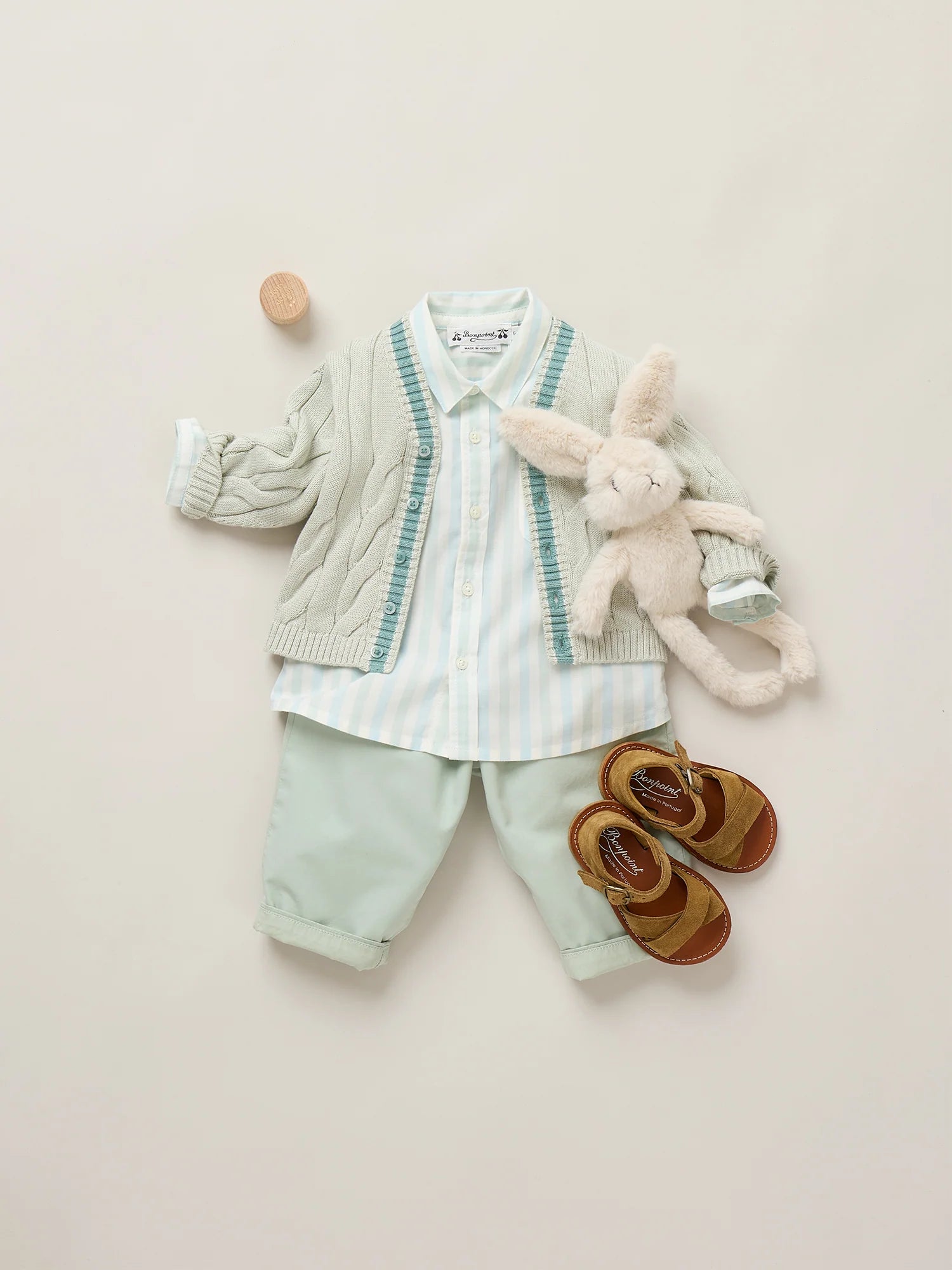 A neatly arranged toddler outfit includes the Bonpoint Baby Boys' Lawson Striped Shirt, a light cardigan, mint pants, brown sandals, a plush bunny toy, and a small round wooden object on a plain background.