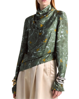 Someone wears the Altuzarra Cara Top, featuring a high-neck design, asymmetrical hem, and signature marbled floral pattern in abstract green. The top's elegant long sleeves are paired with beige pants.