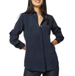 A woman wears the Ann Mashburn Icon Silk Blouse—a long-sleeve, navy blue, collared top with hidden buttons—standing with her hands in her pockets.