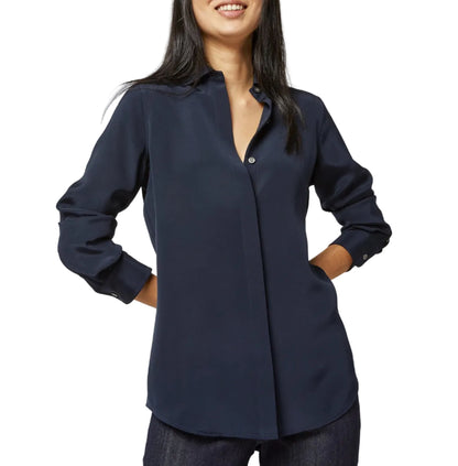 A woman wears the Ann Mashburn Icon Silk Blouse—a long-sleeve, navy blue, collared top with hidden buttons—standing with her hands in her pockets.