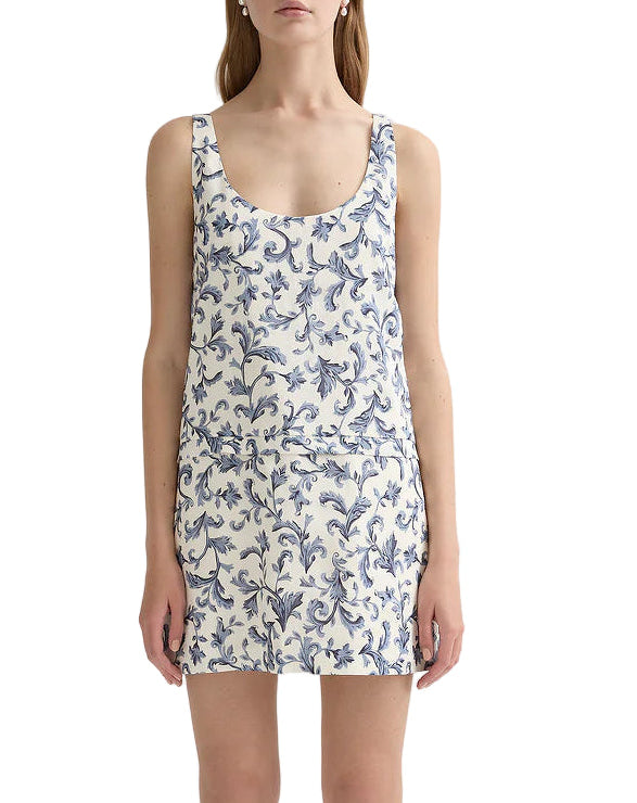 A woman wearing the POSSE Rue Shift Dress—a white linen silk mini dress with a blue floral pattern—and white open-toe shoes, standing against a plain background.