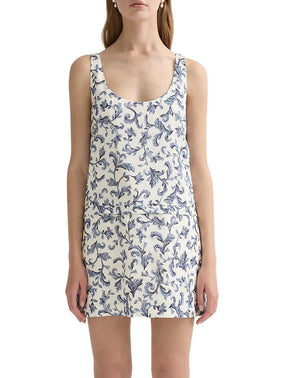 A woman wearing the POSSE Rue Shift Dress—a white linen silk mini dress with a blue floral pattern—and white open-toe shoes, standing against a plain background.