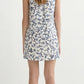 A woman stands facing away, wearing the POSSE Rue Shift Dress with a blue floral pattern and white heels.