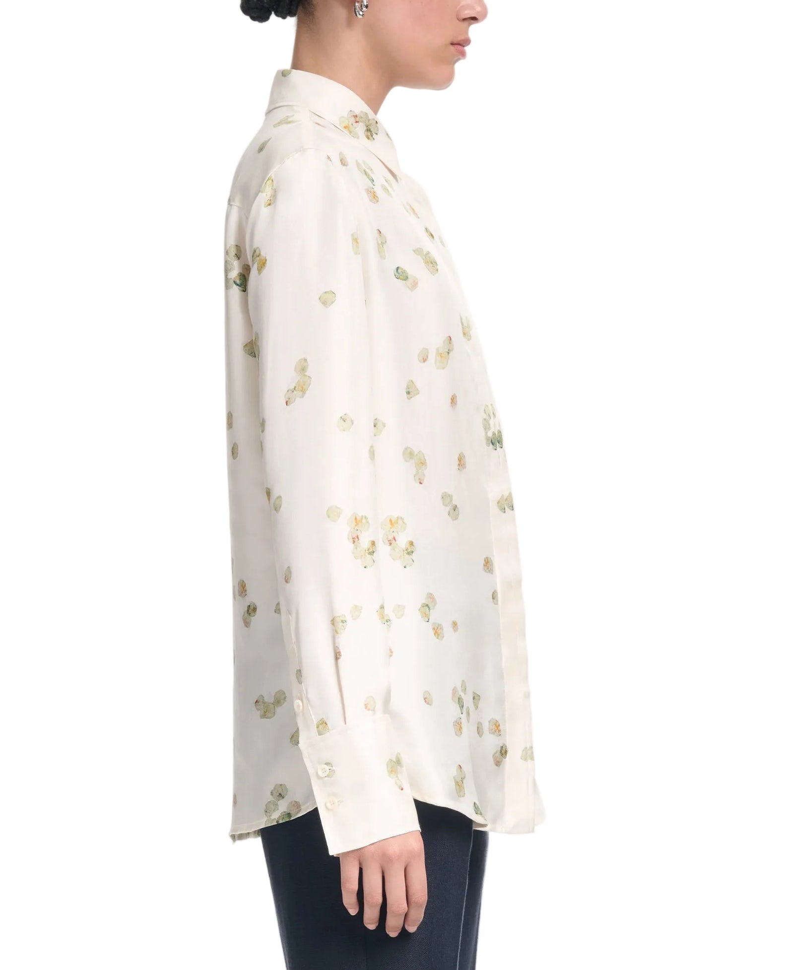A person in profile wears the Altuzarra Chika Top—a relaxed, cream silk blouse with a small floral pattern and long cuffed sleeves—against a plain background.