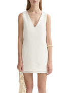 A woman wearing the POSSE Effi Mini Dress—a sleeveless white linen mini dress with a V-neck—paired with a woven bag and a bangle on her right arm.