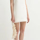 A woman wears the POSSE Effi Mini Dress, a sleeveless white linen mini with a V-neck and adjustable waist tie, paired with a cream crocheted bag and red sandals.