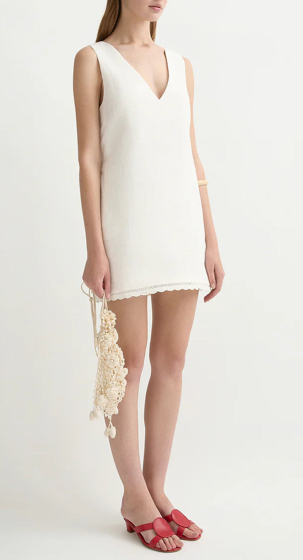 A woman wears the POSSE Effi Mini Dress, a sleeveless white linen mini with a V-neck and adjustable waist tie, paired with a cream crocheted bag and red sandals.