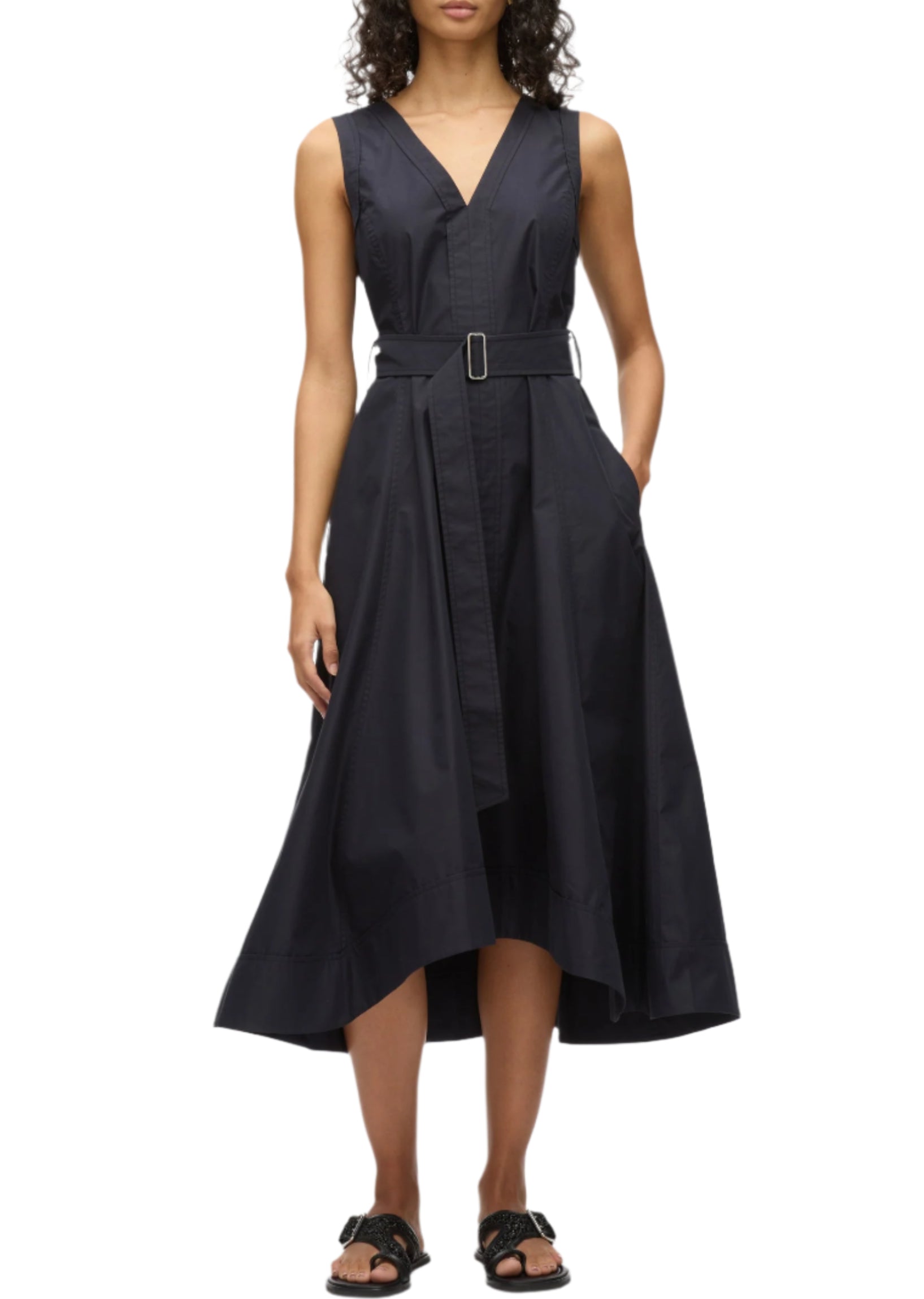 A woman wears the Crisscross Back Midi Dress by 3.1 Phillip Lim—a sleeveless, V-neck style with a black belted A-line skirt in technical poplin, asymmetrical hem, and paired with black sandals.
