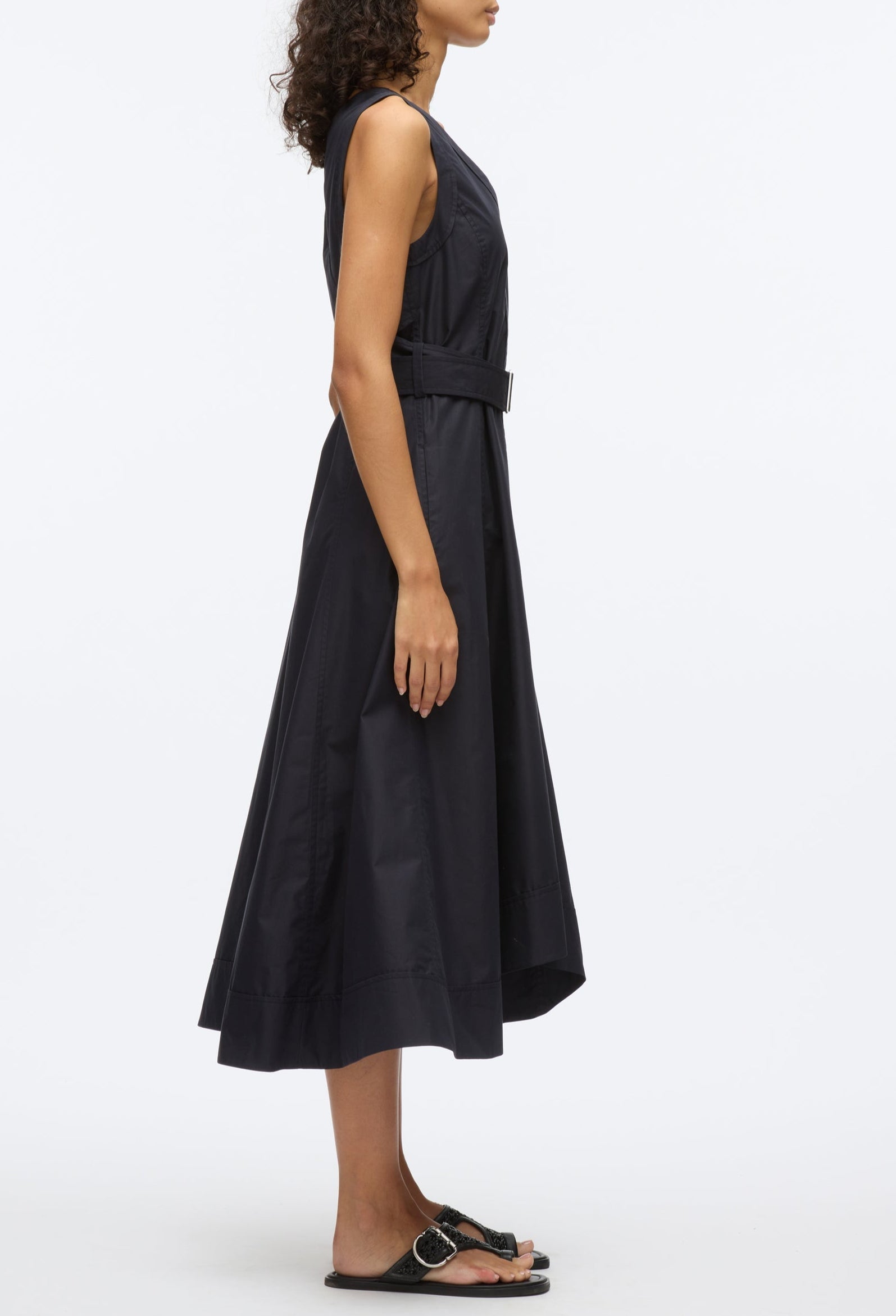 A woman stands in profile wearing the 3.1 Phillip Lim Crisscross Back Midi Dress, a sleeveless black A-line with a belted waist and V-neck, styled with black sandals against a plain white background.
